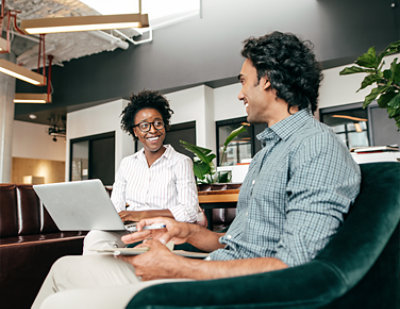 A man seated in a chair looks down at his smartphone, while another person in the background is working on a laptop.