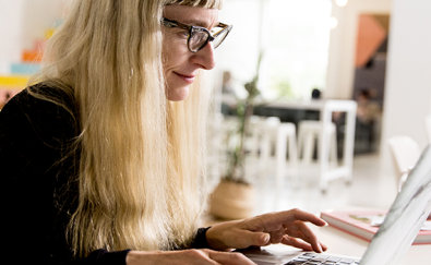 אדם עם שיער בלונדיני ארוך ומשקפיים עובד עם מחשב נישא ליד שולחן בחדר מואר בעיצוב מודרני.