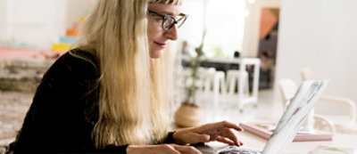 A person with long blonde hair and glasses works on a laptop at a table in a bright room with modern decor.