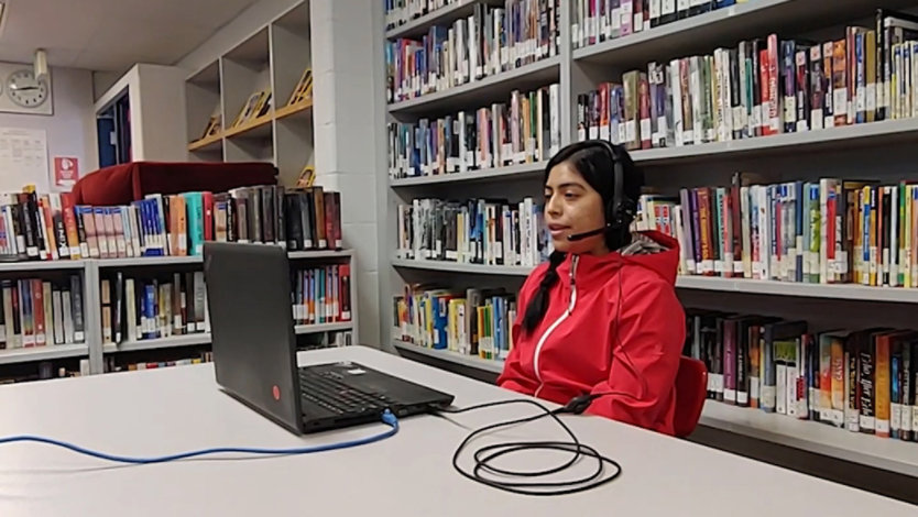 A young female student wearing a red jacket and a headset sitting in a library in front of a laptop.