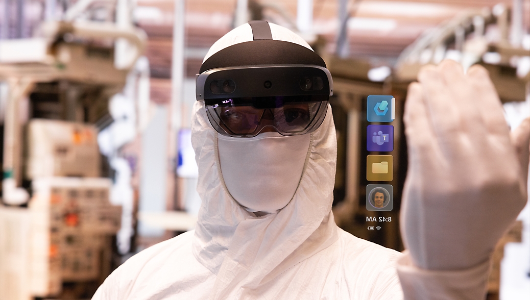 A person in a protective suit and augmented reality headset, interacting with a virtual interface in an industrial setting.