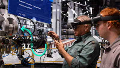 Two engineers, one wearing augmented reality glasses, examine and discuss industrial machinery in a factory setting.