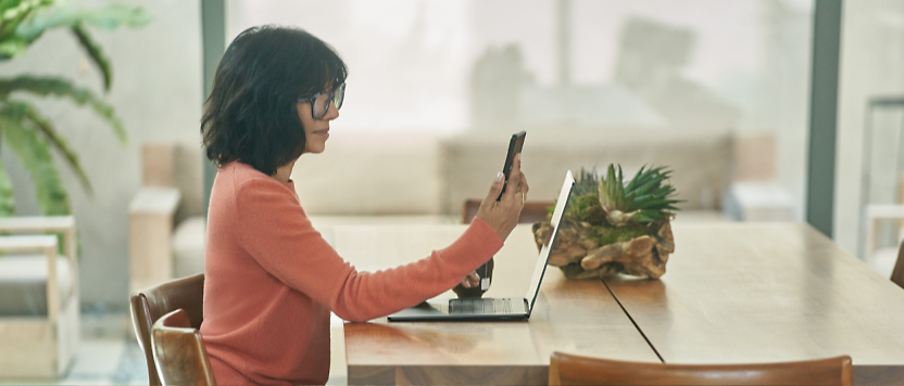 Prillidega naine kasutab sülearvutit ja nutitelefoni tänapäevase puust laua ääres, millel on potitaim 