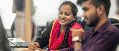 To personer på kontoret som ser på en bærbar datamaskin og har en diskusjon, og én av dem smiler