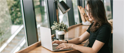 Woman working on a laptop at a modern wooden desk with a beverage beside her.