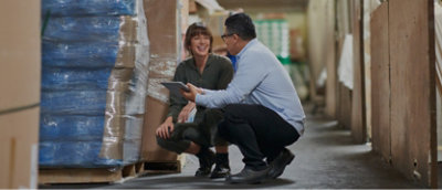 Two people kneeling in a warehouse looking at a tablet.
