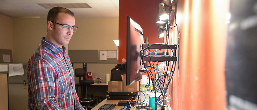 Um homem de óculos e camisa axadrezada concentra-se no ecrã de um computador num ambiente de escritório desorganizado.