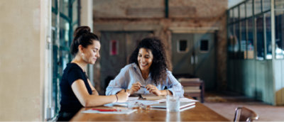 Zwei Frauen sitzen an einem Holztisch in einem gut beleuchteten Raum mit großen Fenstern, diskutieren über Dokumente und lächeln.