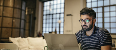 A person wearing glasses and a striped shirt is sitting in front of a laptop in a room with large windows.
