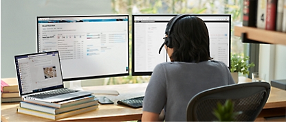 En kvinna sitter vid ett skrivbord med två bildskärmar, en bärbar dator ovanpå en hög med böcker och ett headset.