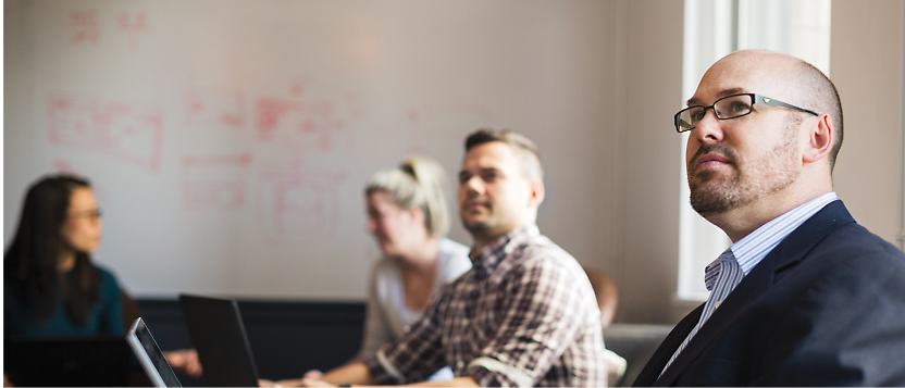 Forretningsfolk i et mødelokale med én fokuseret person i forgrunden og et whiteboard med noter i baggrunden.