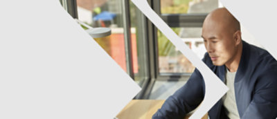 A bald man in a blue sweater sits at a table, looking intently at a laptop in a brightly lit modern cafe.