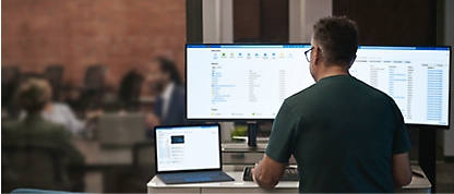En mand, der sidder ved et skrivebord med to skærme og en bærbar computer foran sig.