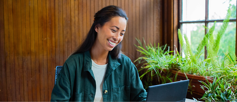 Woman smiling while using a laptop in a room with plants and wooden paneling.