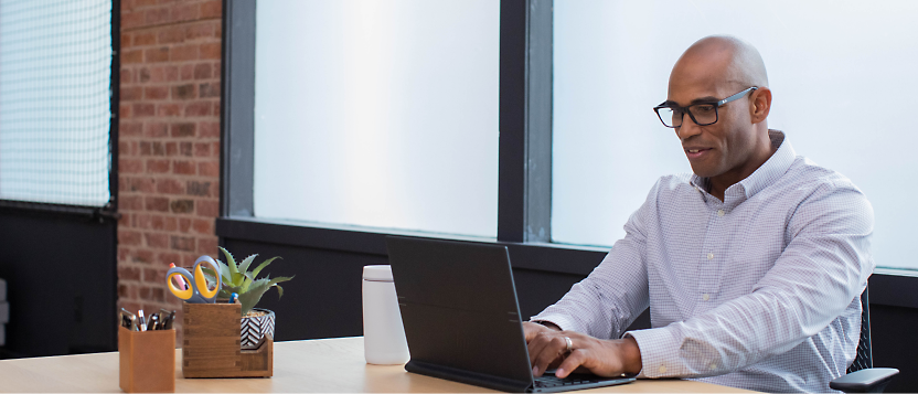 Seorang pria mengenakan kacamata dan kemeja berkerah yang bekerja di laptop di lingkungan kantor modern.