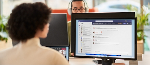 Uma mulher a colaborar uma reunião do Microsoft Teams enquanto trabalha num escritório aberto num monitor duplo