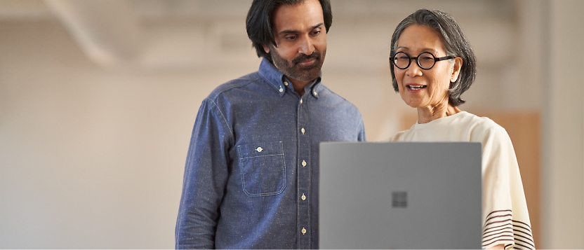 Twee personen staan naast elkaar en kijken naar een laptopscherm. De ene draagt een blauw shirt en de andere draagt een bril.