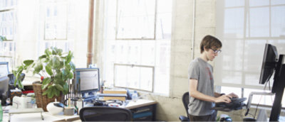 Birden fazla bilgisayar ekranı ve masasının bulunduğu aydınlık bir ofis alanında ayakta duran bir masa kullanan adam.