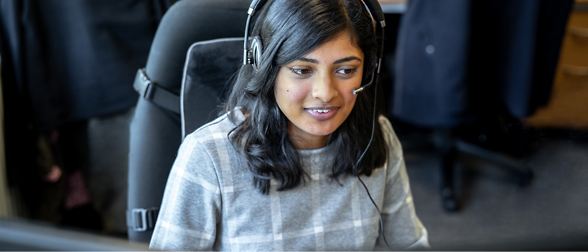 A woman wearing headphones and a microphone smiles while working at a busy office environment.