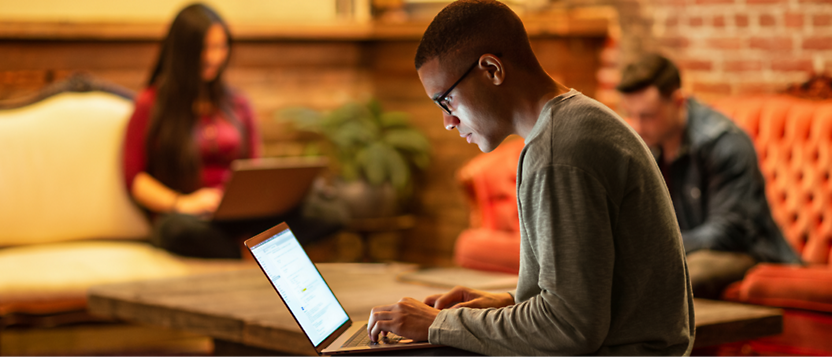 Een jonge man met een bril die een laptop gebruikt in een informele werkruimte met andere mensen op de achtergrond.