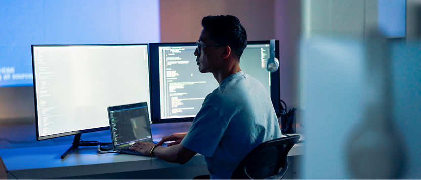 A man in glasses working on multiple computer screens with code displayed in a dimly lit office.