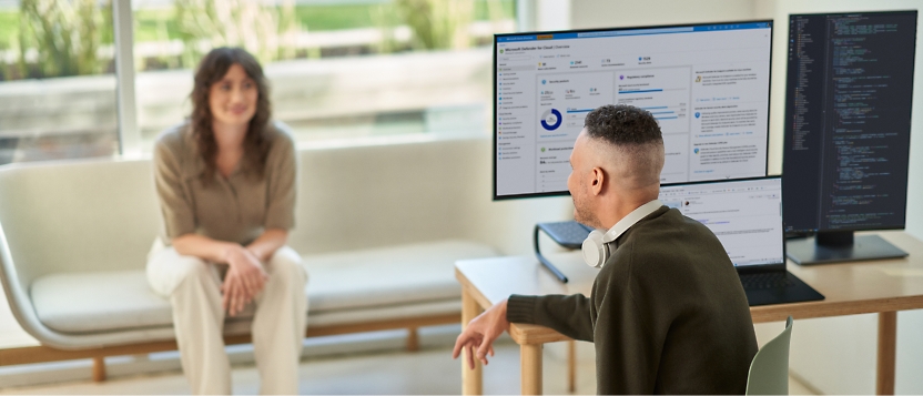 Dos personas que participan en una conversación en un área de trabajo con portátiles con varias pantallas que muestran datos y código.