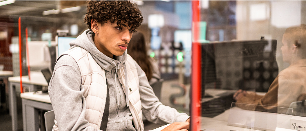 Young man with curly hair sitting at a computer desk in an office, looking away thoughtfully.