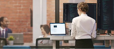 Uma mulher está de pé numa secretária com dois monitores à sua frente.