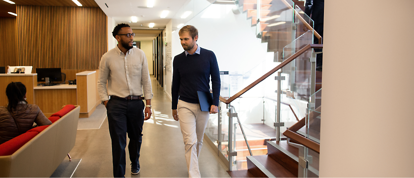 Dwóch mężczyzn spacerujących i rozmawiających w nowoczesnym korytarzu biurowym.