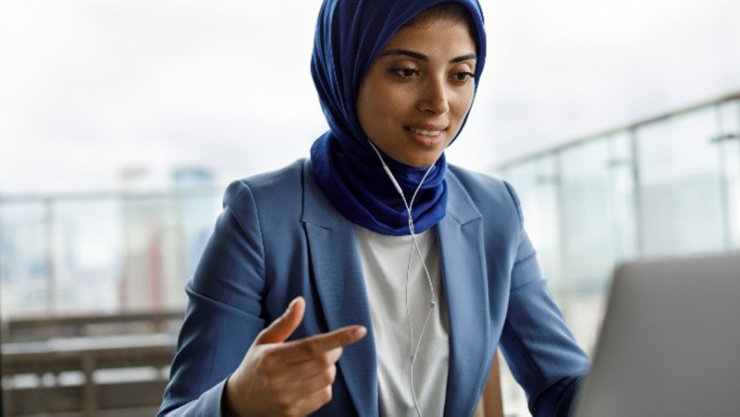 Young woman with blue headscarf, blazer, and headphones working on laptop