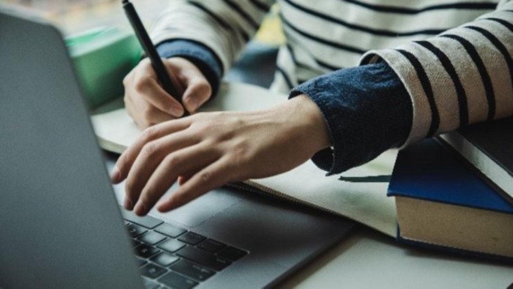 Seated person typing on laptop and writing in notepad, with elbow resting on stack of books