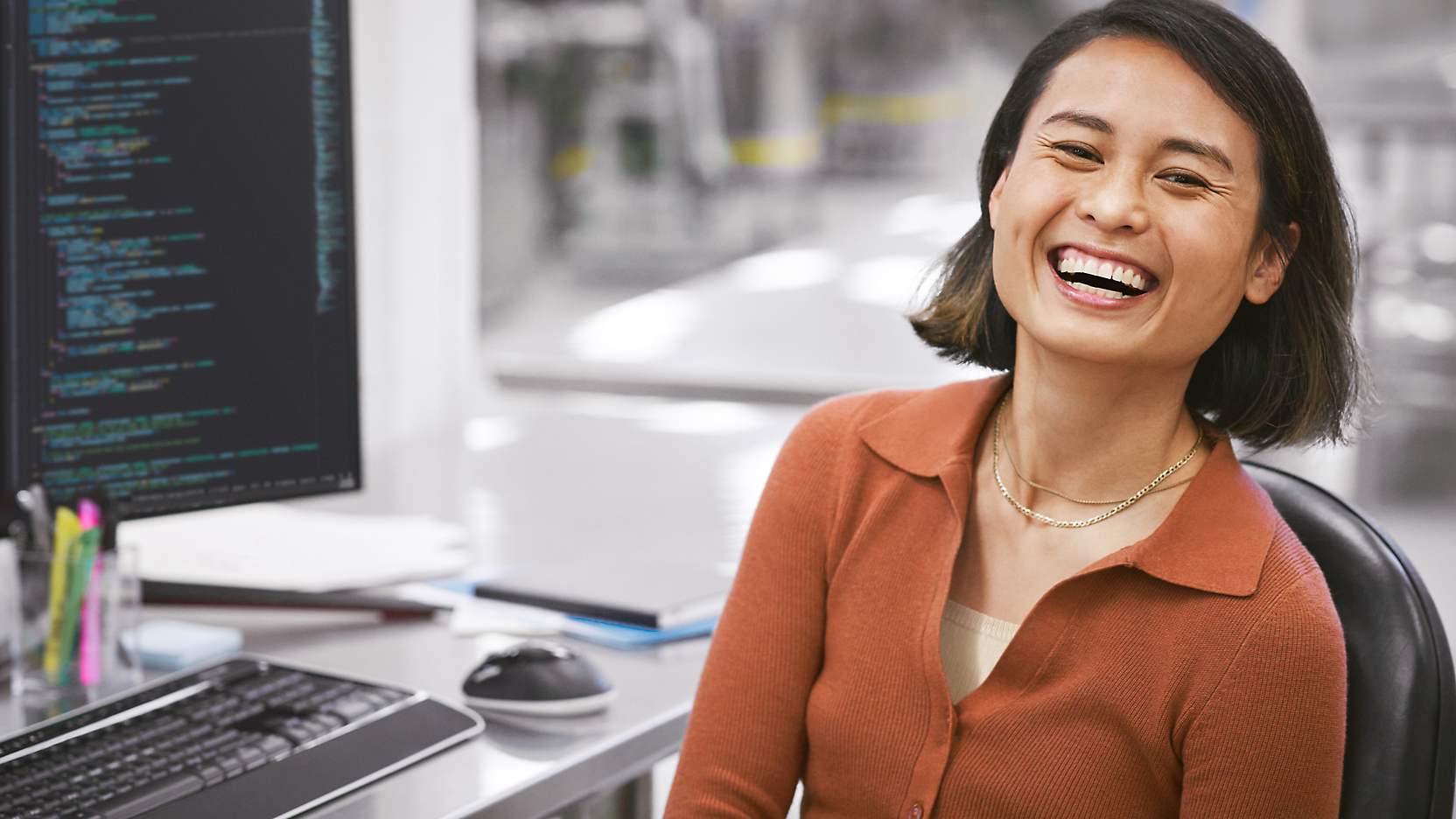 Una donna felice seduta a un tavolo con un computer che mostra codice sullo schermo in un ufficio moderno.
