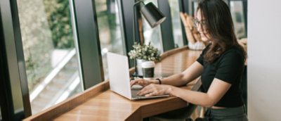 Woman working on laptop at a wooden desk near a window.