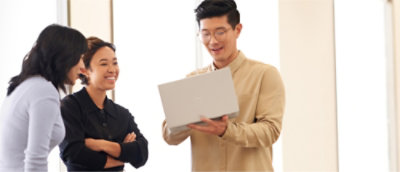 Three people are gathered, looking at a laptop held by a man in a beige shirt.