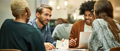 Four people are seated at a table, engaged in conversation, with a laptop and a book in front.