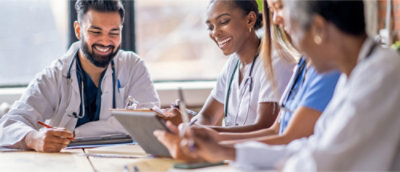 A group of healthcare professionals in white coats and scrubs sit at a table, smiling and discussing notes.