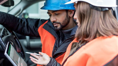 Zwei Kollegen in einem Auto mit Sicherheitswesten und Schutzhelmen, die auf ein Tablet schauen