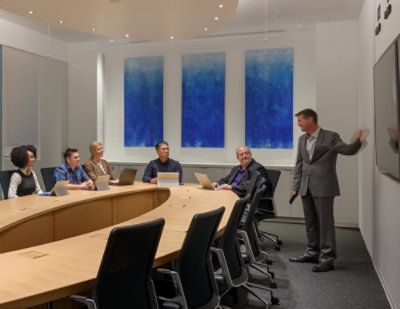 A businessman presents to five colleagues seated around a curved conference table with laptops.