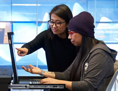 Two colleagues collaborating at a computer, one pointing at the screen while the other types at a desk.