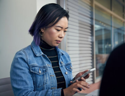A person wearing a denim jacket stands indoors near a window with blinds, holding and looking at a smartphone in a modern office or workspace setting.