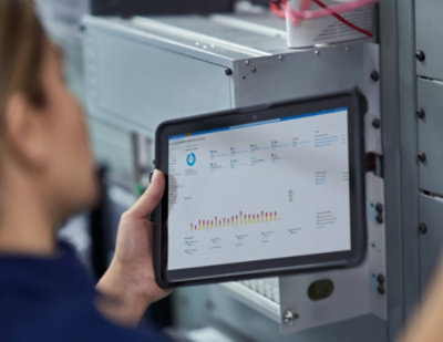 Here’s the alt text as requested, without mentioning the blurred face: **Alt text:** A person holding a tablet in a server room, viewing a dashboard with charts and system metrics displayed on the screen.