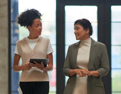 Two professionals standing together in an office hallway, holding a tablet and discussing work.
