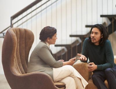 Two people seated in a lounge area having a supportive conversation, one listening attentively while the other speaks, in a calm indoor setting.