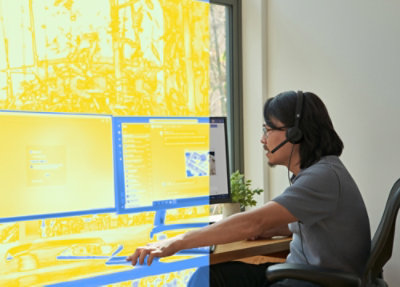 Person working at a desk with dual monitors, wearing a headset in a bright workspace.