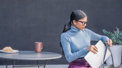 Here’s a concise, neutral alt text that **does not mention the blurred face**: **Alt text:** A person sitting at a small round table outdoors, holding an open notebook, with a mug and plate on the table against a plain gray wall.