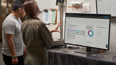 Two people standing at a workspace, reviewing analytics dashboards on a large monitor in a workshop-style office setting.