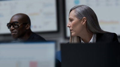 Person working at computer desk in office, colleague seated nearby reviewing content on screen