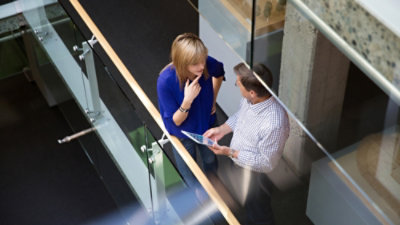 Two people standing in a modern office hallway, looking at a tablet together, seen from above.