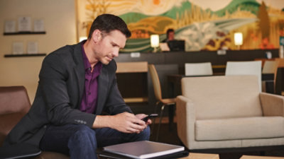 Person sitting in a lounge area wearing a blazer, looking at a smartphone with chairs and a mural in the background.