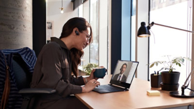  Person sitting at a desk holding a mug and participating in a video call on a laptop, with a desk lamp and plant nearby.